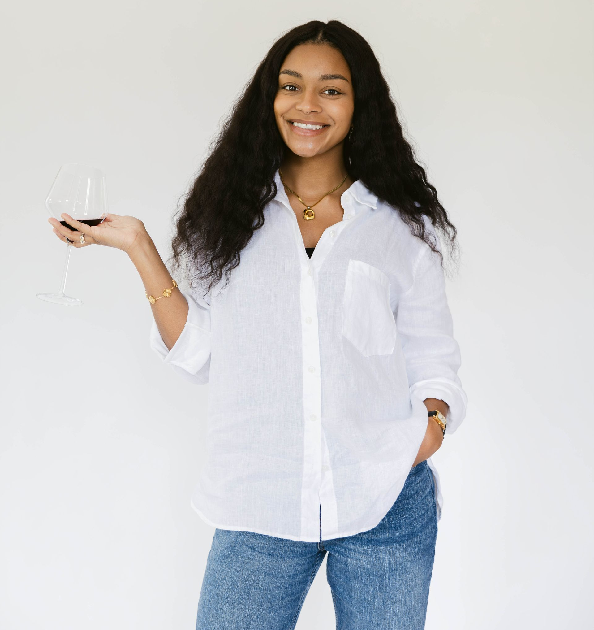 Woman standing against a white wall, holding a wine glass and looking to the smiling forward.