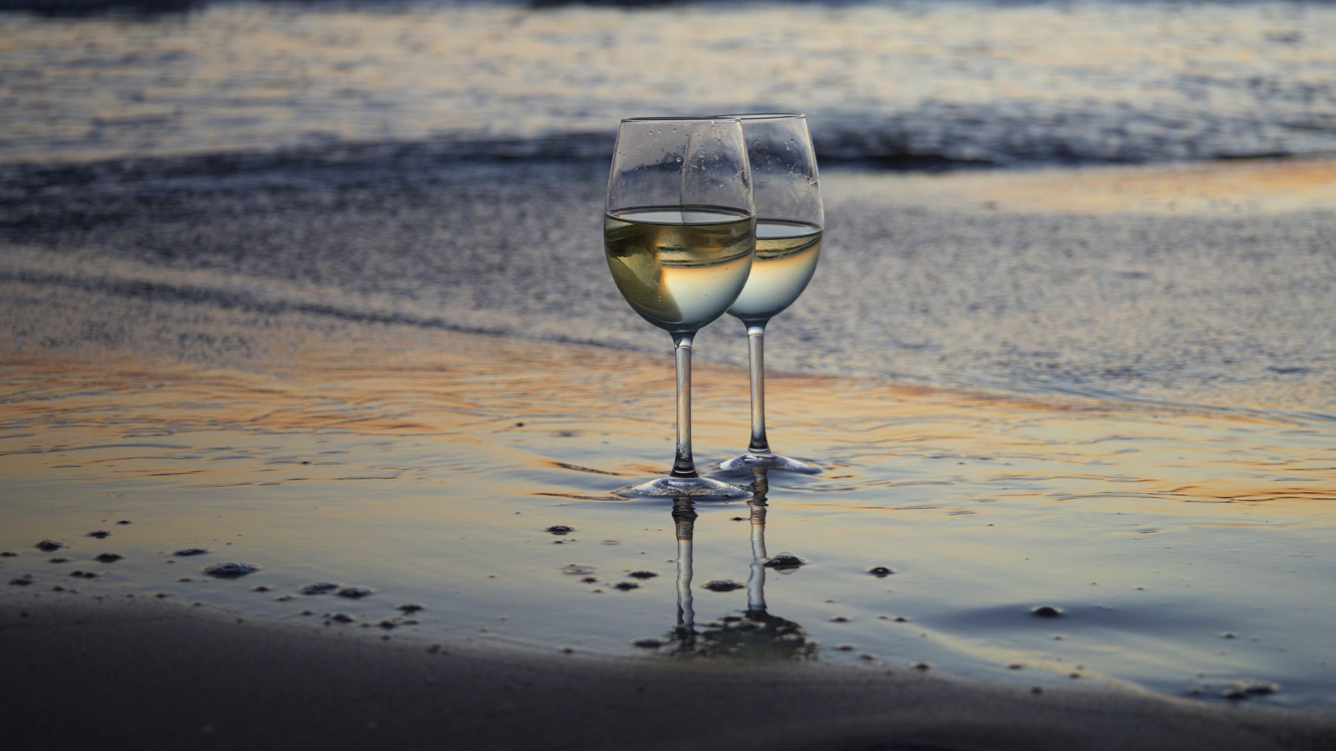 two wine glasses sitting on the sand near a body of water