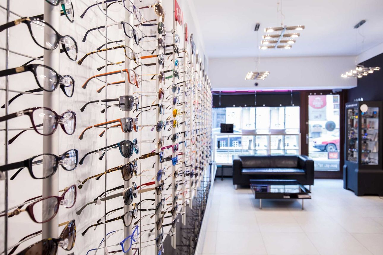 Eyeglass display wall in a bright optical shop with a black waiting bench and front window. Eyeglasses displayed on wall racks in a bright optical store with a seating area near the entrance