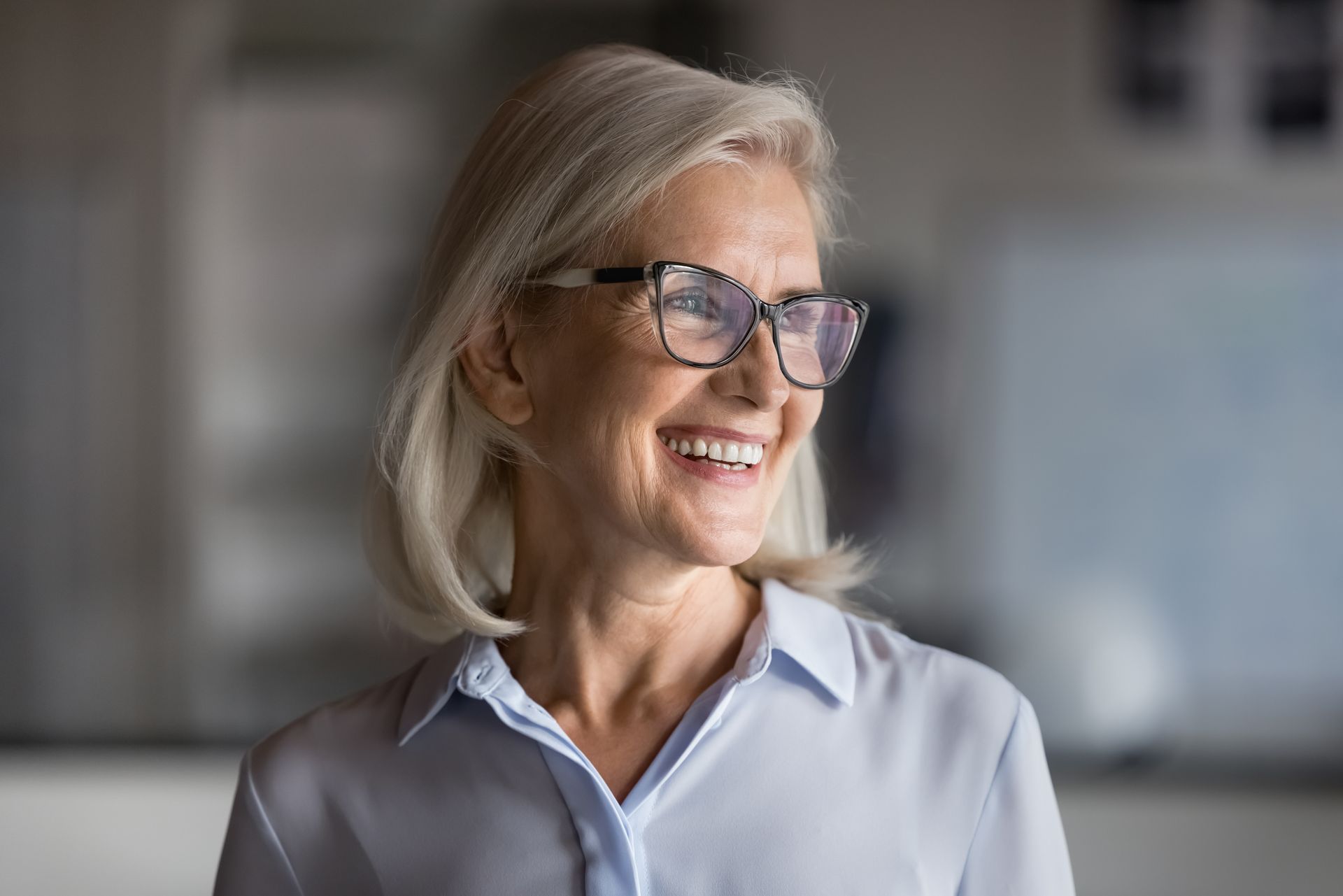 Smiling woman with glasses in a light blue shirt, looking to the side indoors