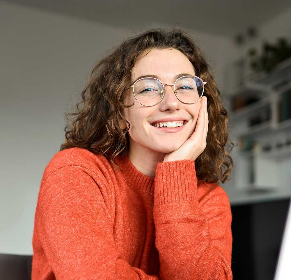 Smiling person in glasses and an orange sweater resting their chin on their hand indoors