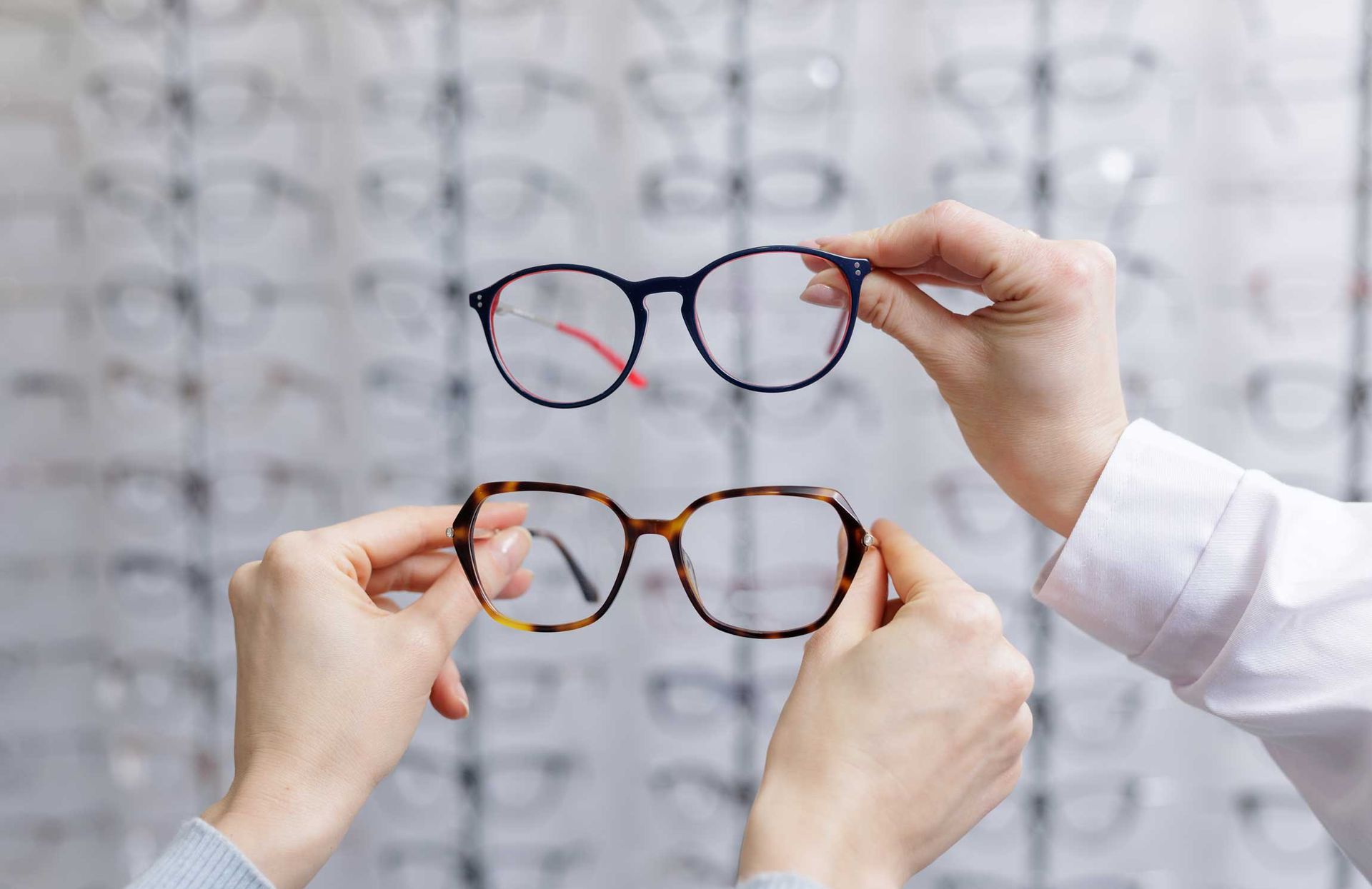 Hands holding two eyeglasses frames up to a lens display wall
