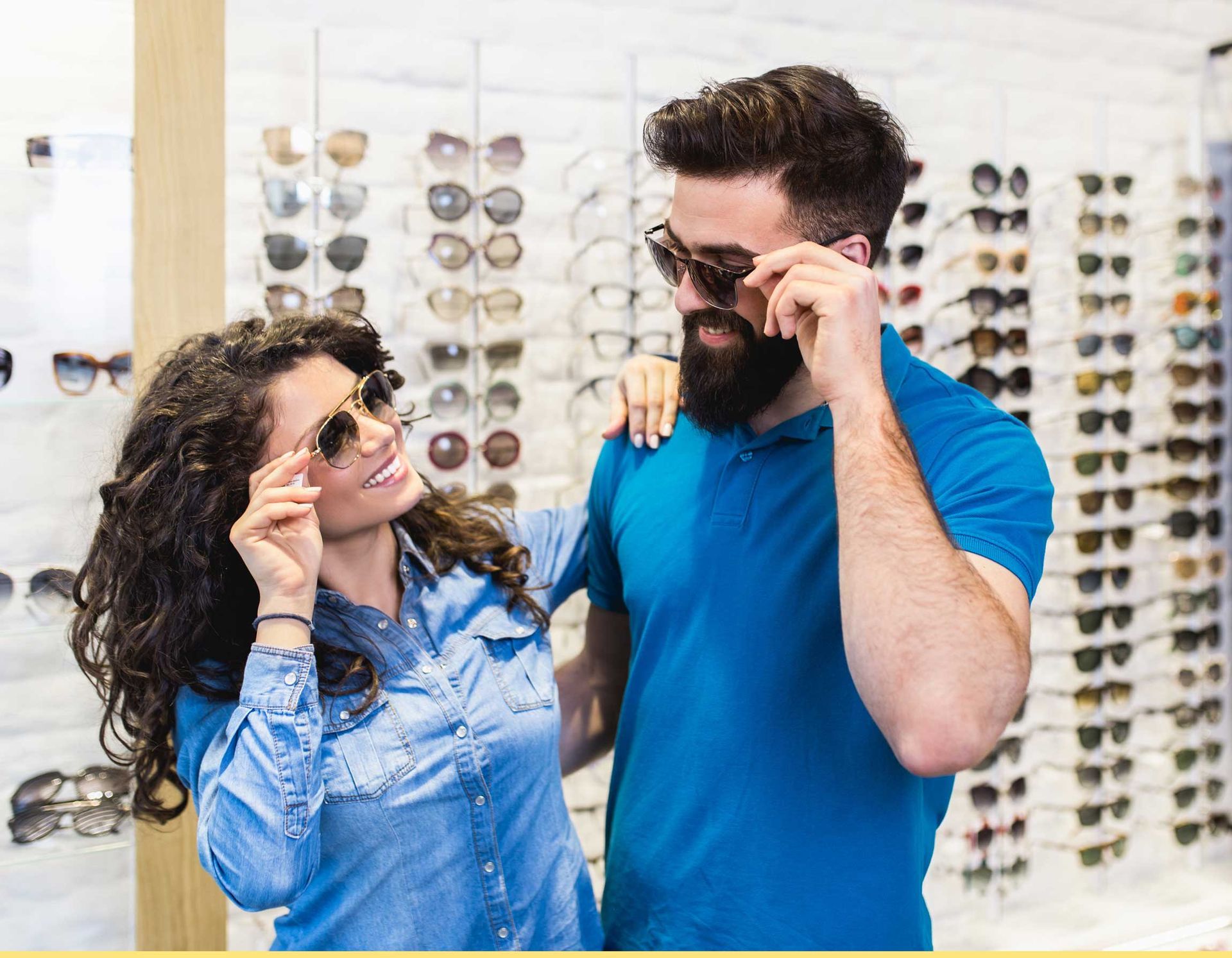 Two people trying on sunglasses in an eyewear store, with rows of glasses in the background.