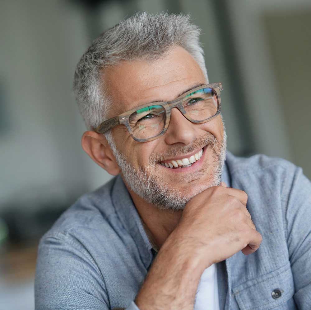 Smiling man with gray hair and glasses, resting his chin on his hand indoors