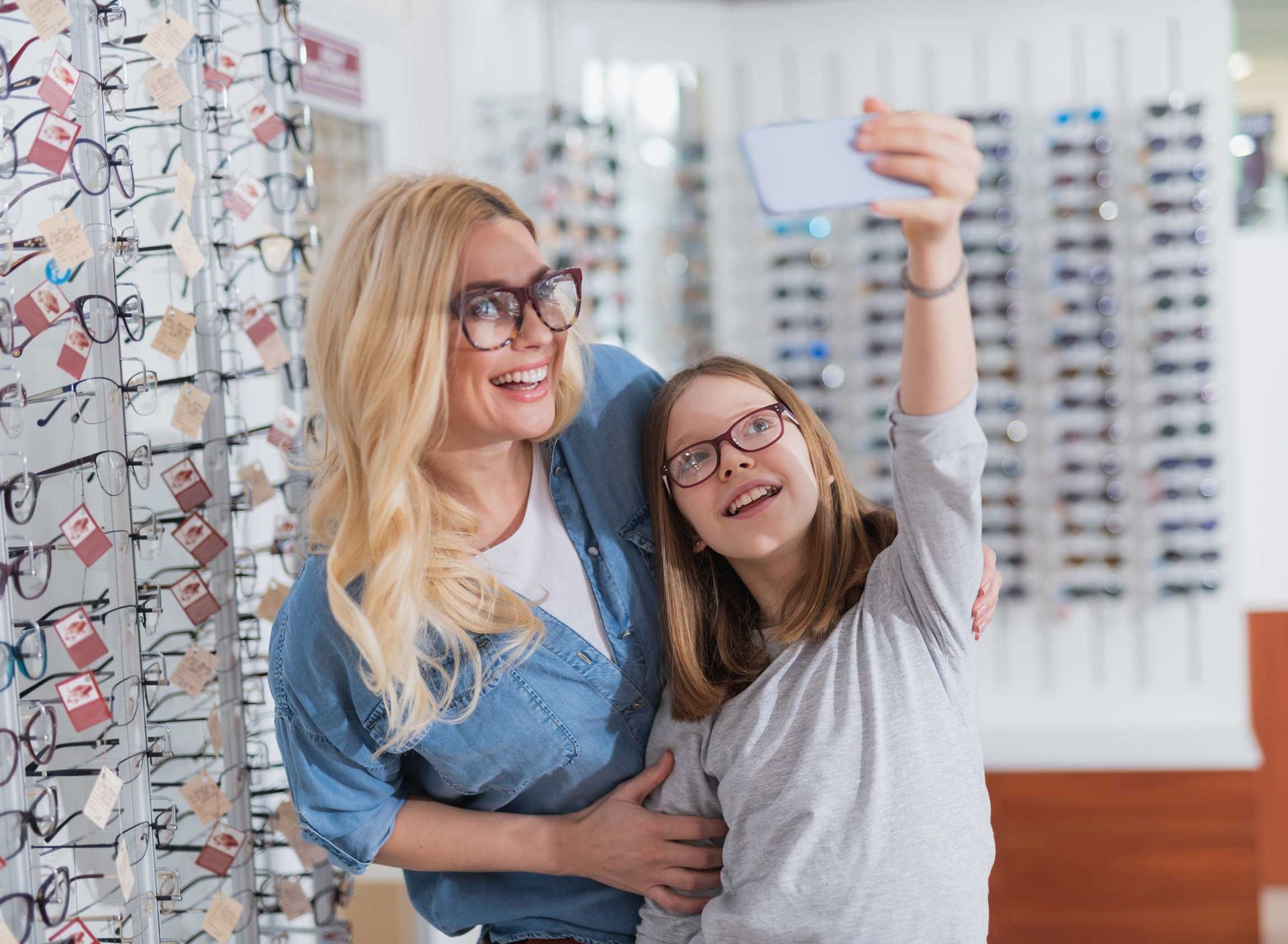 Two people smiling in an eyeglasses store, one taking a selfie in front of rows of frames