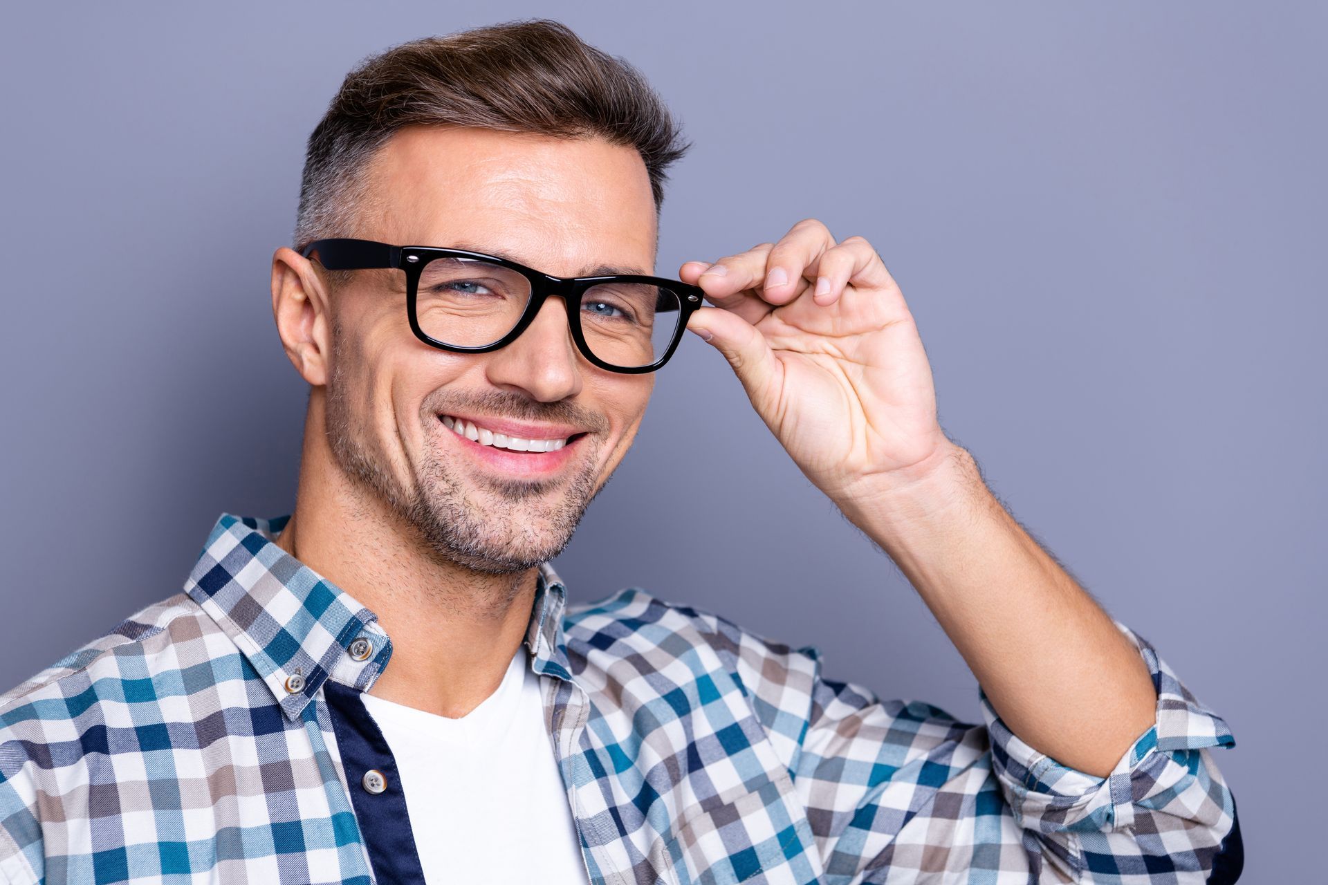 Smiling man adjusting black glasses, wearing a blue plaid shirt against a gray background