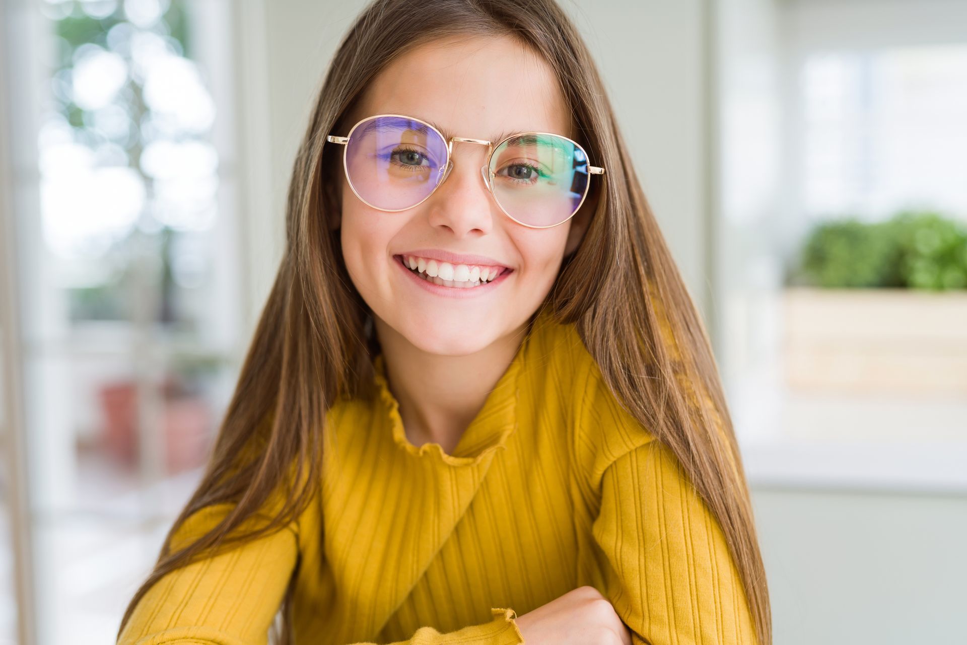 Smiling person in a yellow sweater and glasses indoors, leaning forward with arms crossed