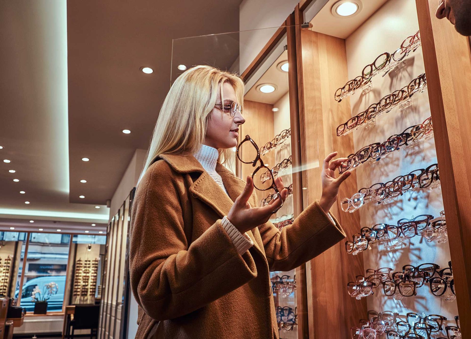 Woman trying on eyeglasses in a stylish eyewear store, browsing frames on a display wall.