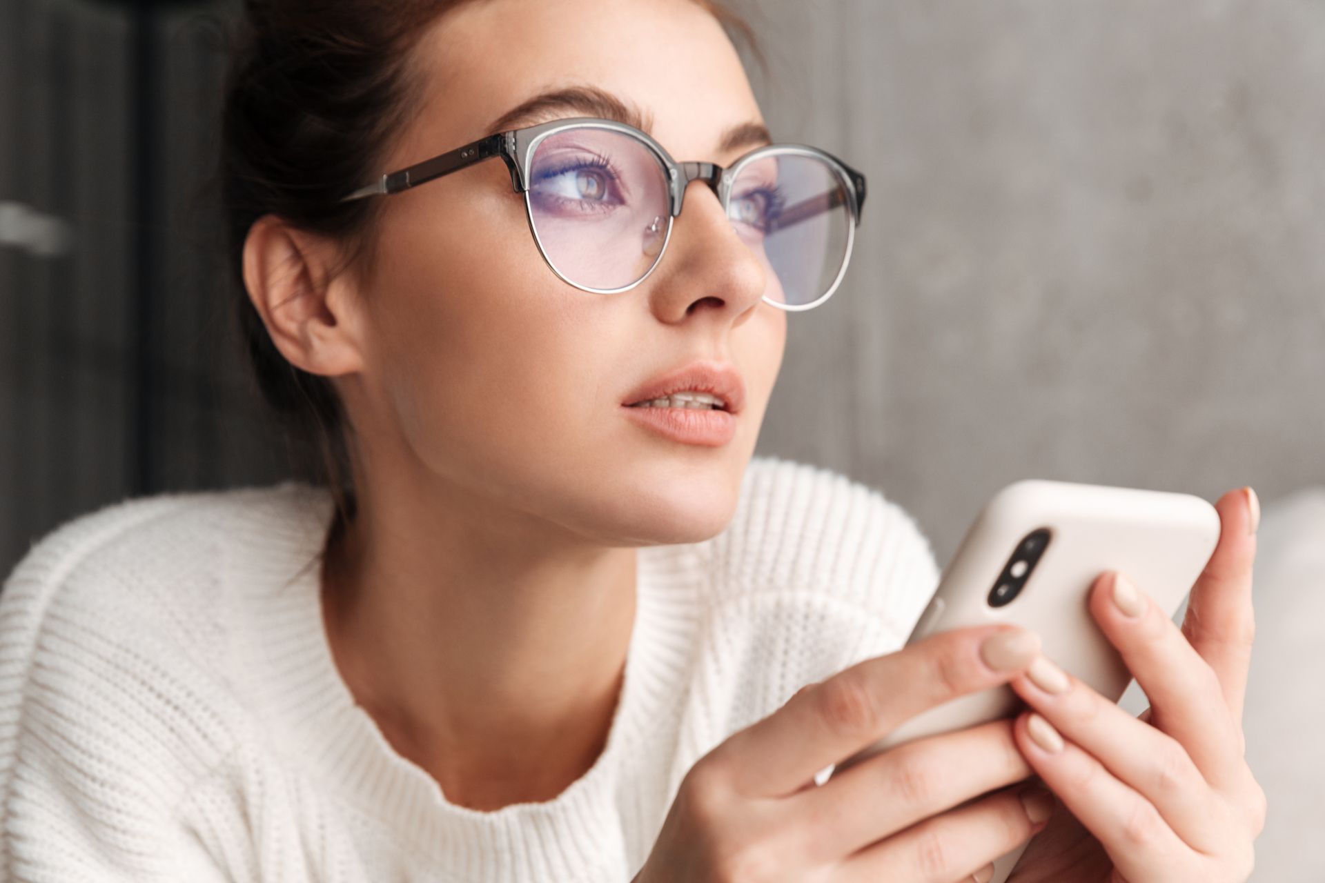 Woman in glasses holding a smartphone and looking thoughtfully to the side