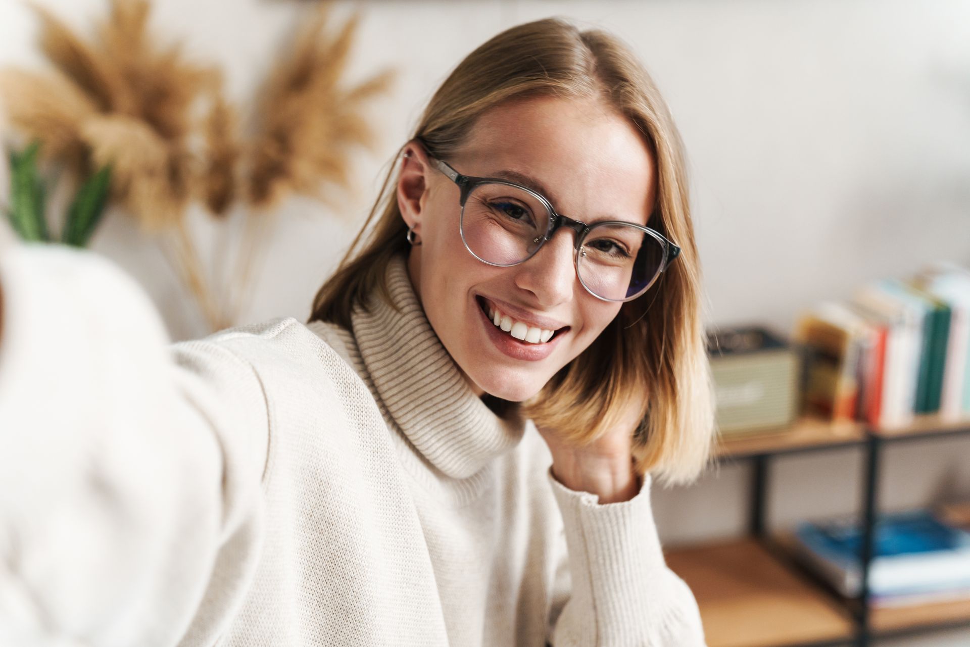 Smiling person with glasses taking a selfie in a bright room with a plant and bookshelf behind them