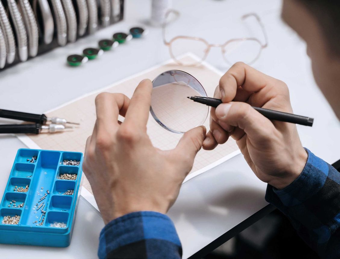 Hands examining a round watch crystal with a marker, beside watch parts and tools on a workbench Hands examining a round eyeglass lens with a pen at a jeweler’s workbench, tools nearby