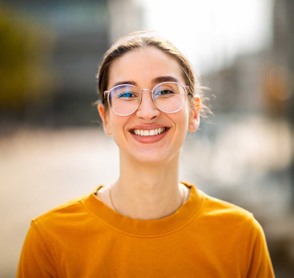 Smiling person in glasses and an orange shirt, standing outdoors with a blurred city background