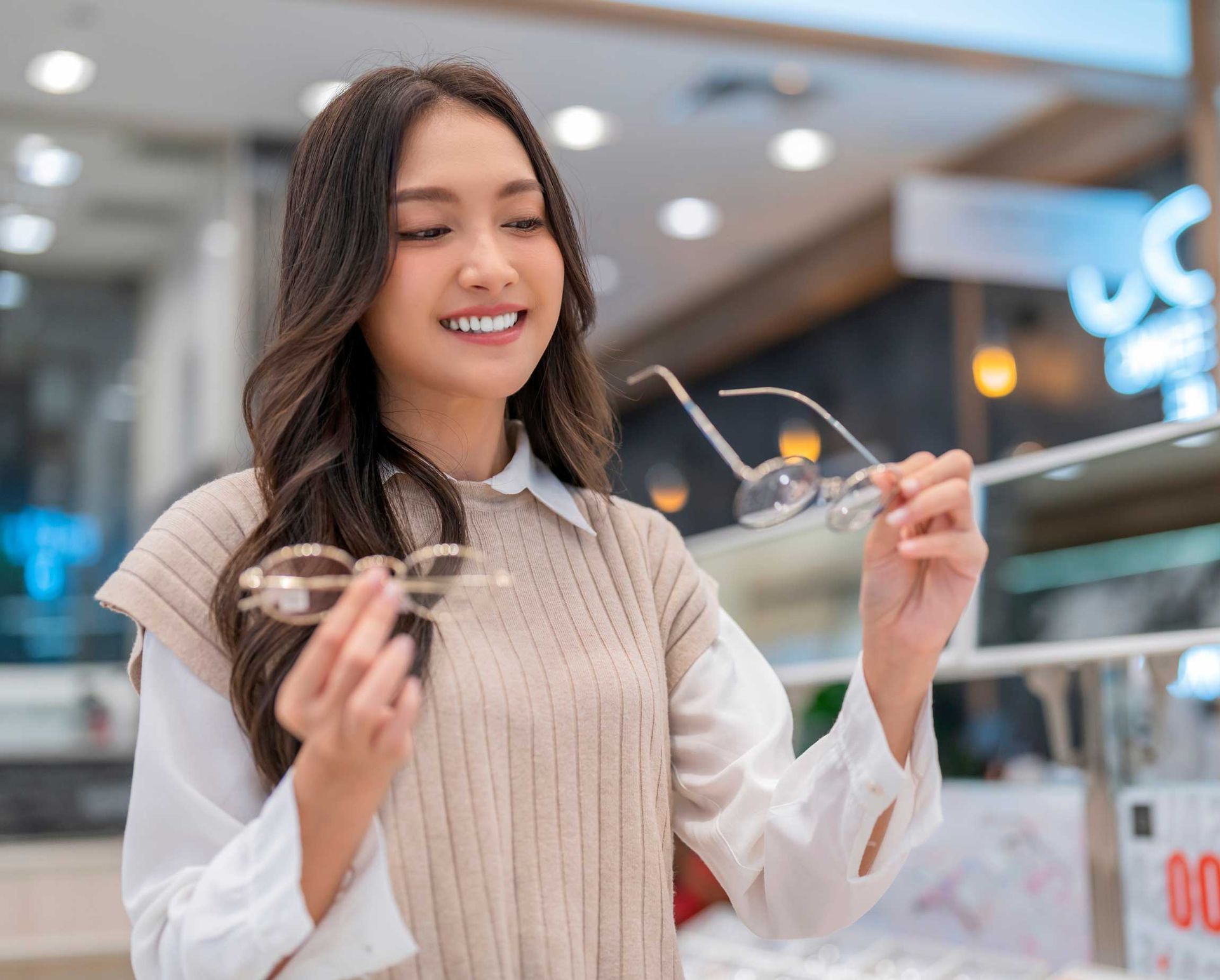 Woman smiling while holding eyeglasses in a bright shopping mall and looking at them