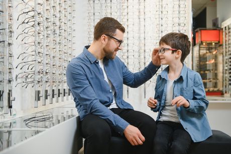 Two people are talking in an optical shop, seated beside rows of eyeglasses. Two people talking in a workshop, seated by rows of eyeglasses and equipment, one in denim jacket and one in glasses