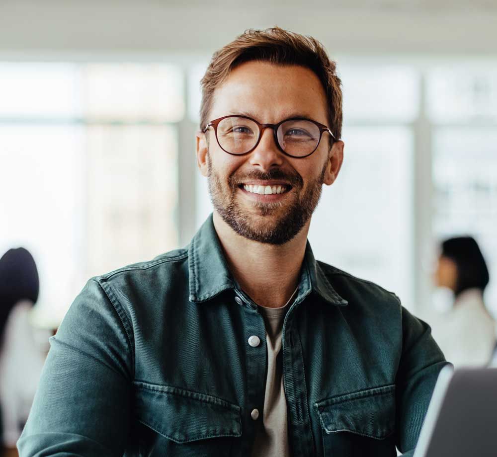 Smiling man with glasses in a denim jacket, sitting in a bright office with a laptop