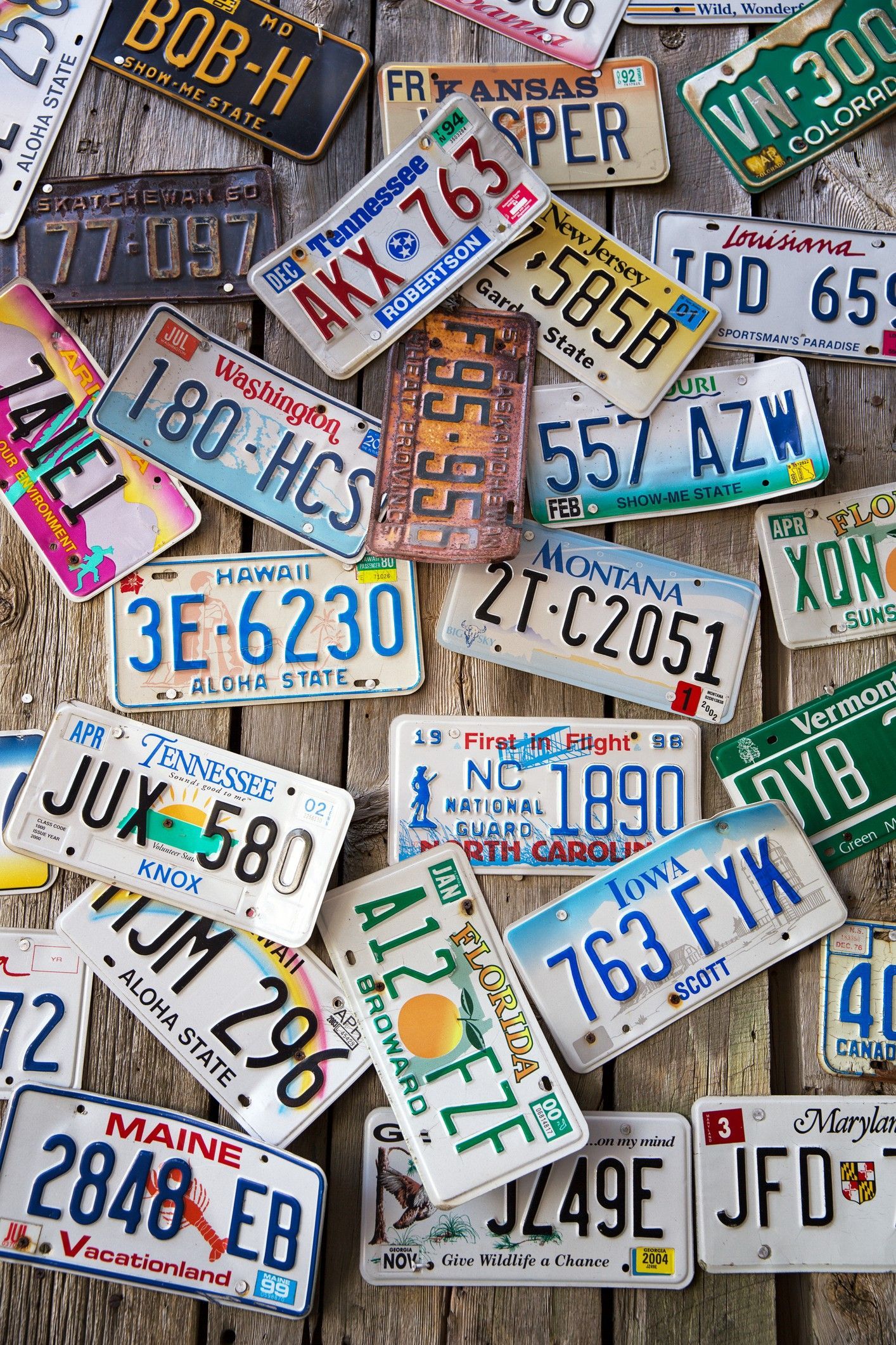 Scattered collection of license plates, various states and designs, on a weathered wooden surface.
