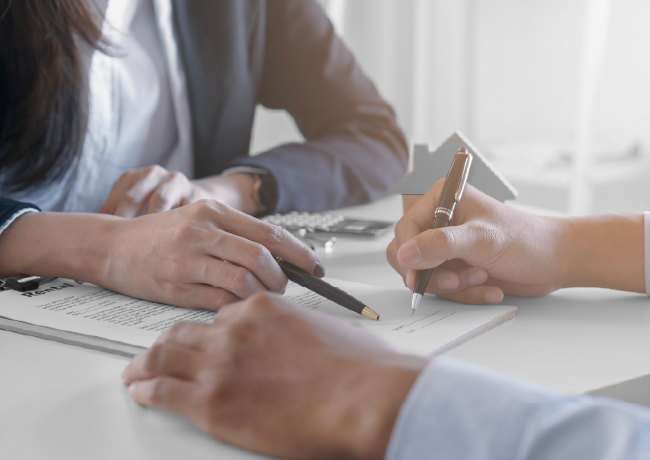 Hands signing a document, likely a contract, with a pen, over a desk with a model house.