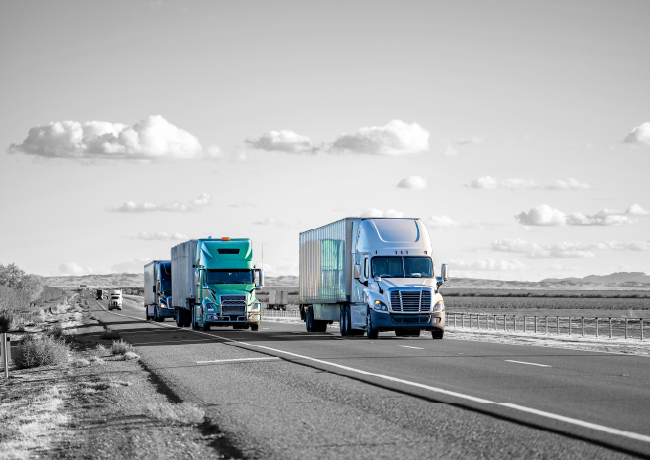 Semi-trucks, teal and silver, on a highway under a cloudy sky. Black and white background.