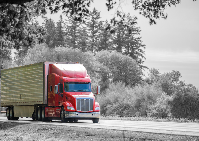 Red semi-truck driving on a road with a black and white background of trees.
