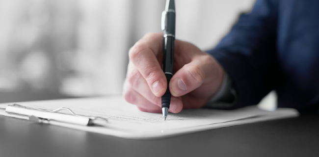 A person's hand holding a pen, writing on a clipboard with a stack of papers.