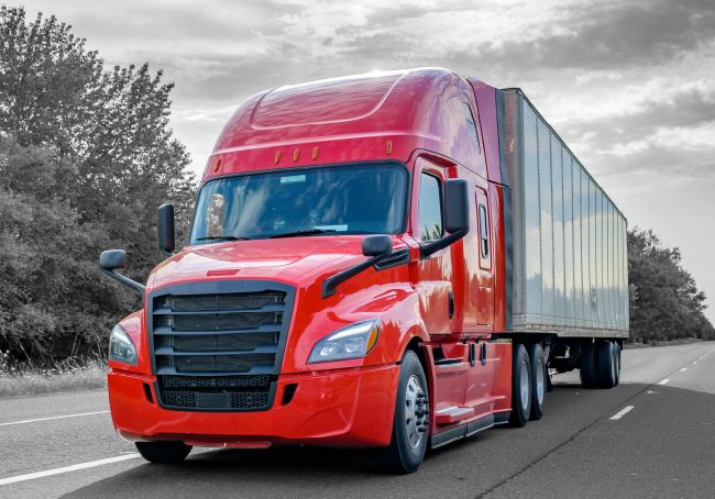 Red semi-truck on a highway, hauling a trailer, overcast sky in the background.