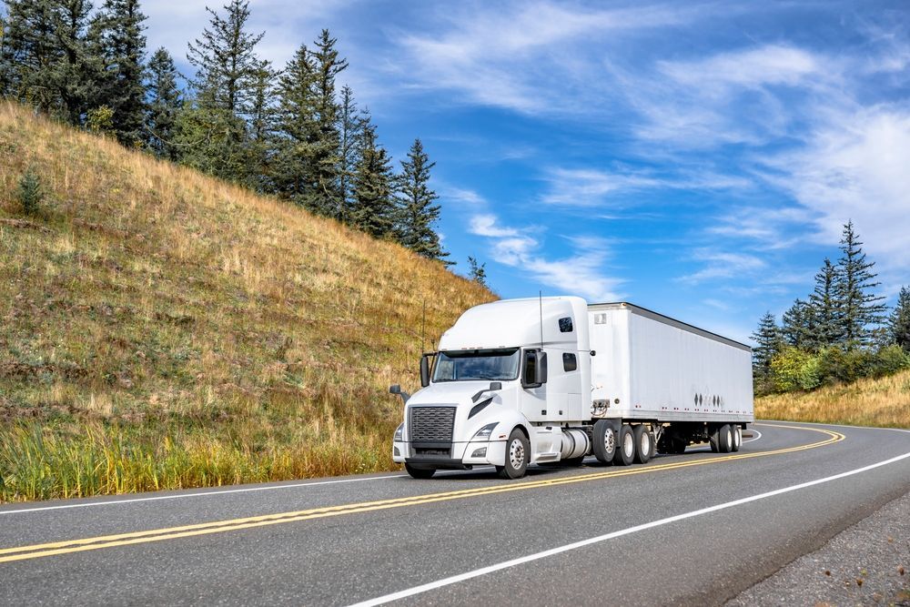 White semi-truck driving on a curving highway beside a grassy hillside and pine trees under a blue sky