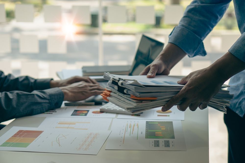 Two people work together at a desk, reviewing a stack of paperwork and printed financial charts.