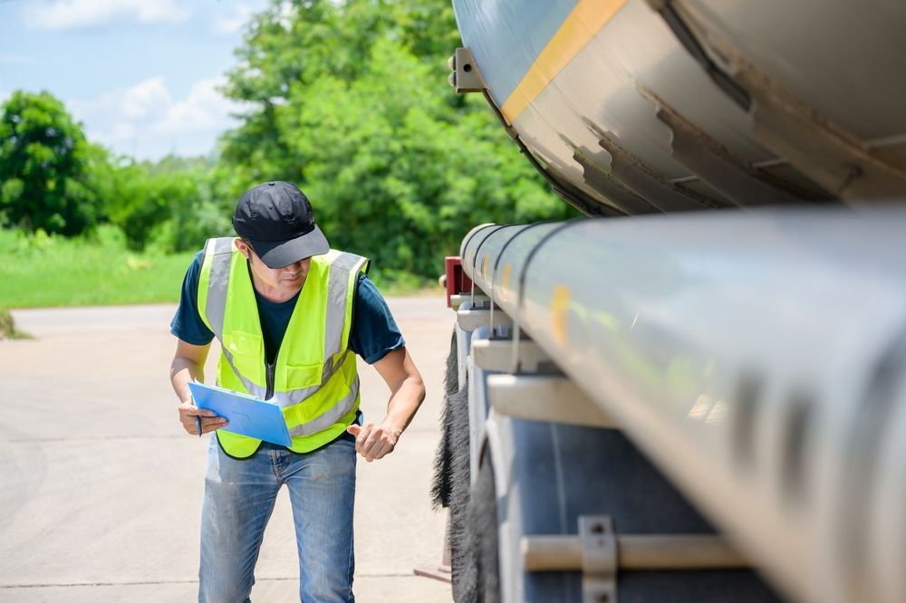 Worker in a reflective vest inspecting a tanker truck outdoors on a sunny roadside