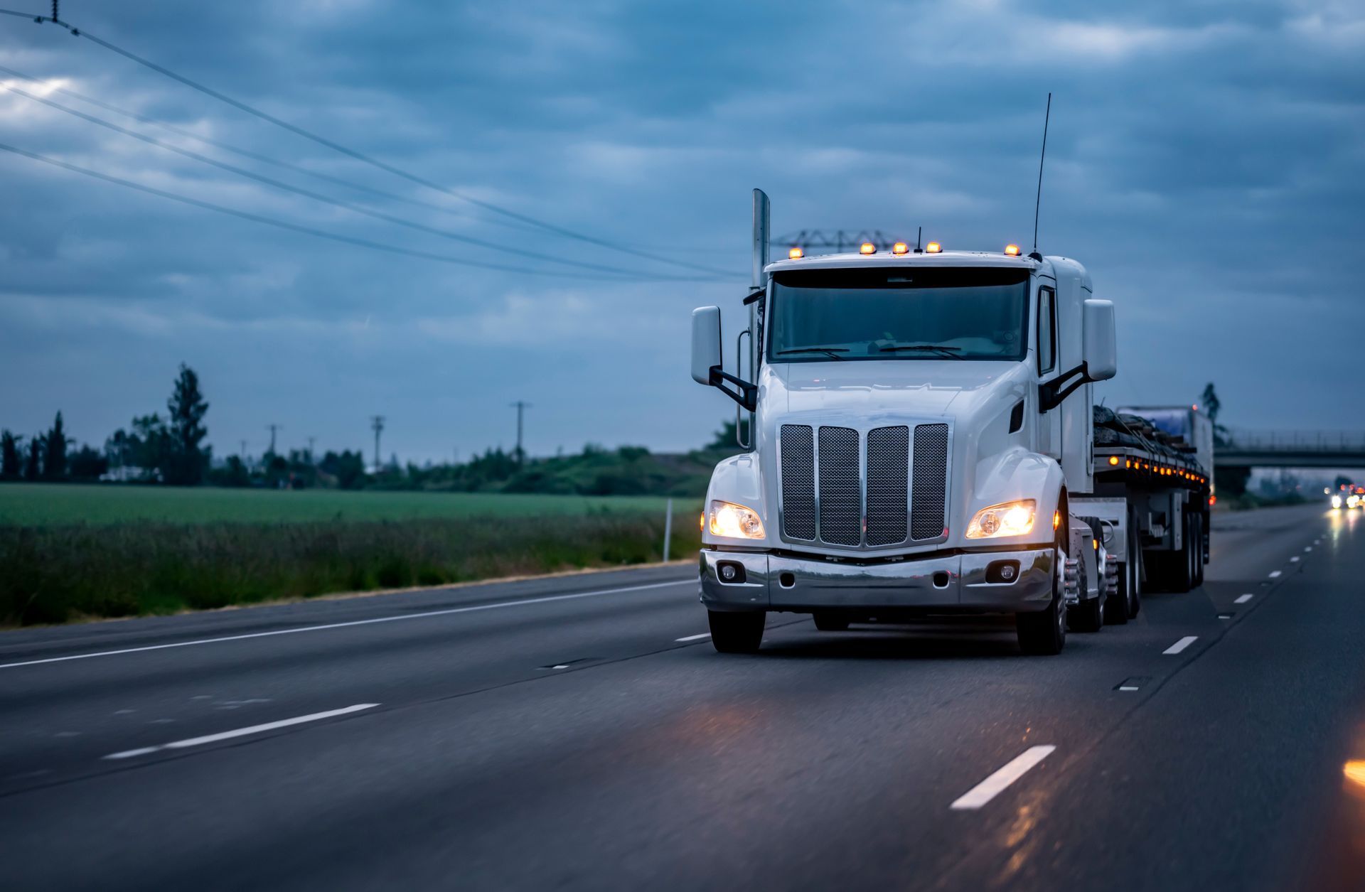 White semi-truck driving on a highway at dusk, with cloudy sky and fields in the background.