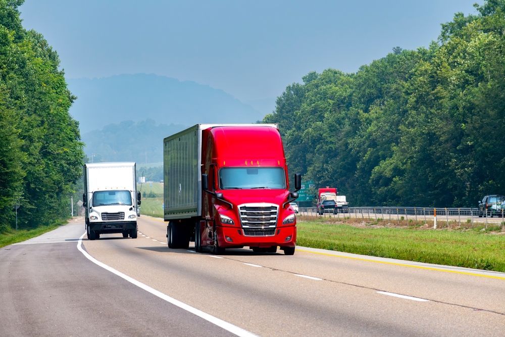 Two semi-trucks on a highway, a red one in front and a white one behind. Green trees and a hazy mountain are in the background.