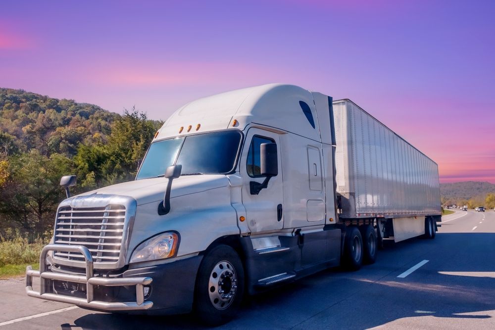 Semi-truck driving on a road with a forest and colorful sky in the background.