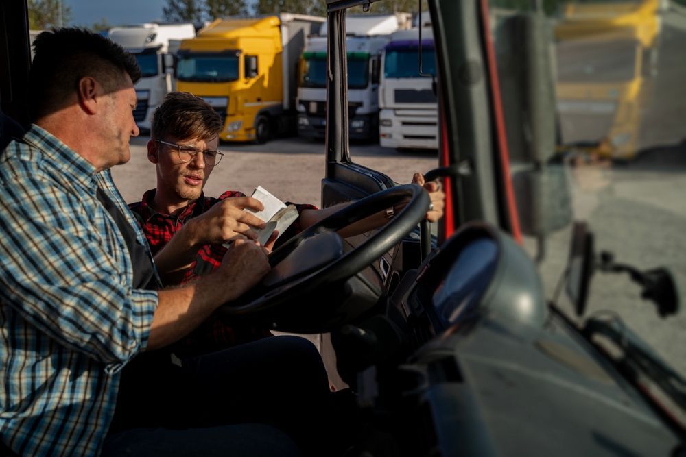 Man and young person in a truck, looking at a paper. Trucks are parked in the background.