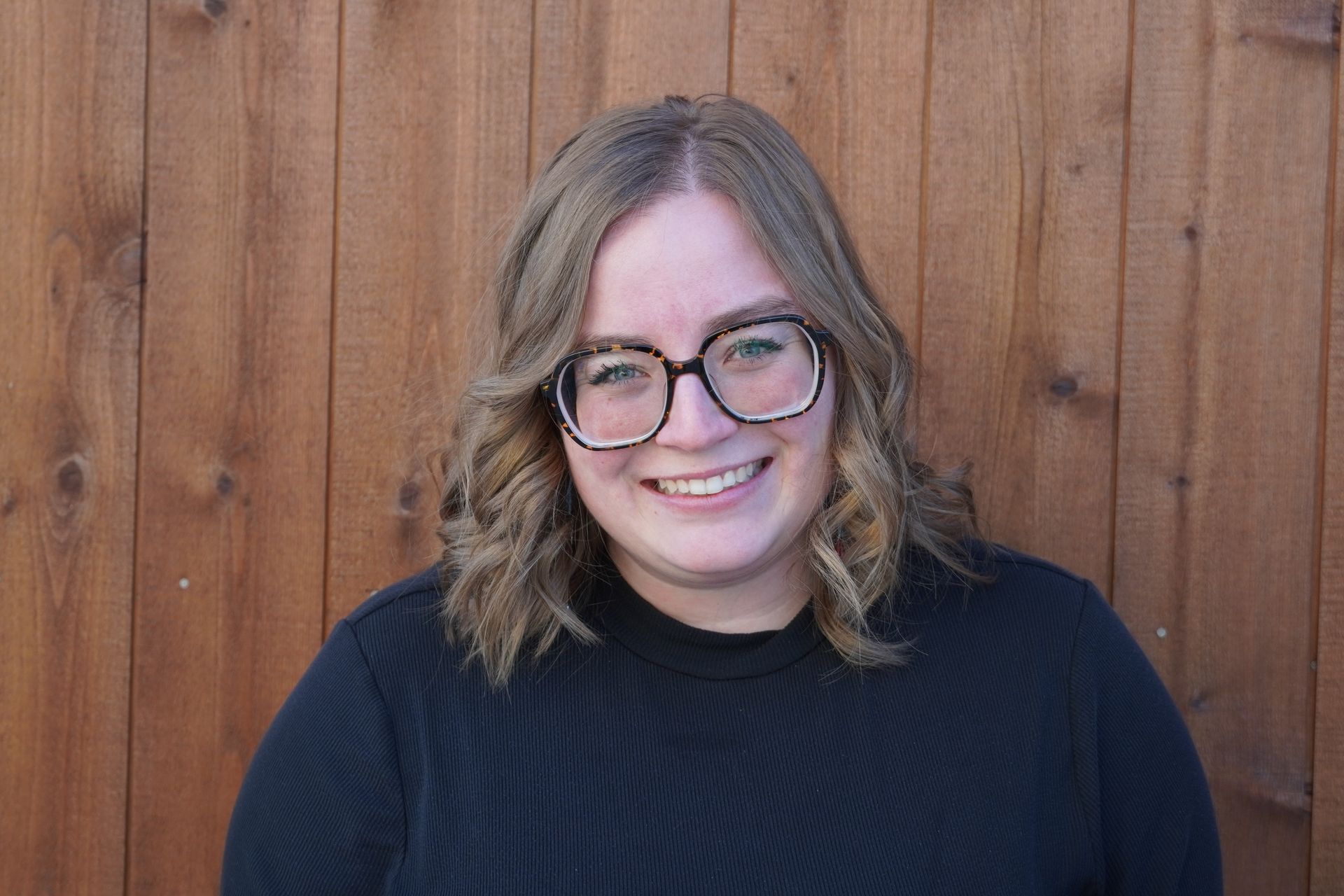 a woman wearing glasses and a black shirt is smiling in front of a wooden fence .