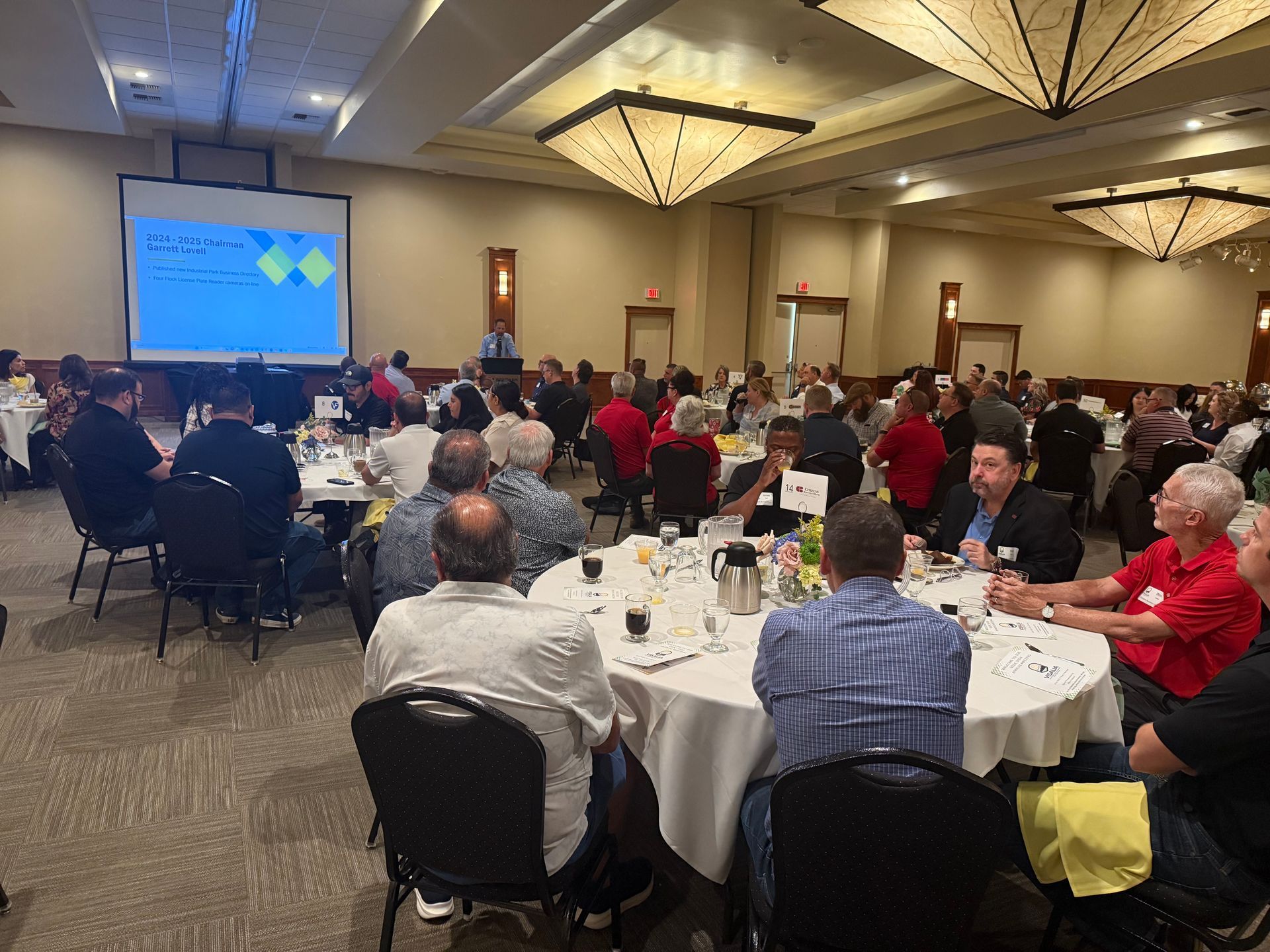 Large room with round tables, attendees, and a presentation on a screen.