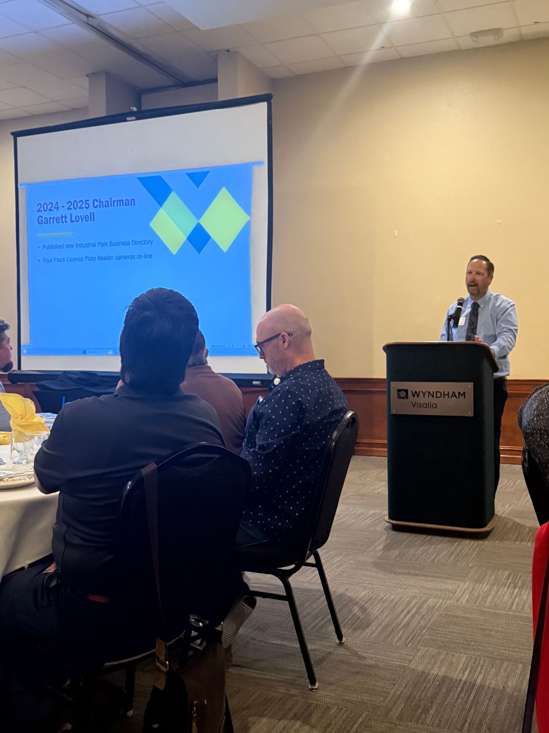 Man giving a presentation at a podium, screen in background with a blue and yellow graphic. Audience seated.