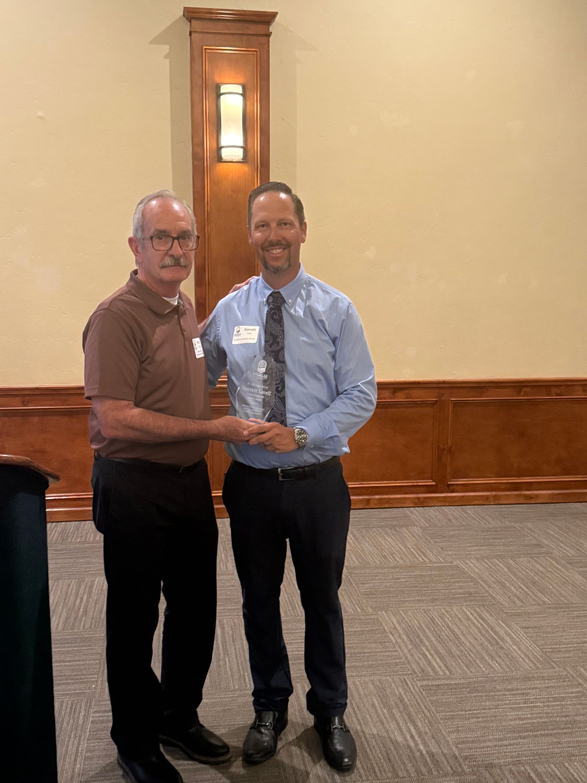 Two men shaking hands indoors at an event. One in brown shirt, the other in blue, holding an award.