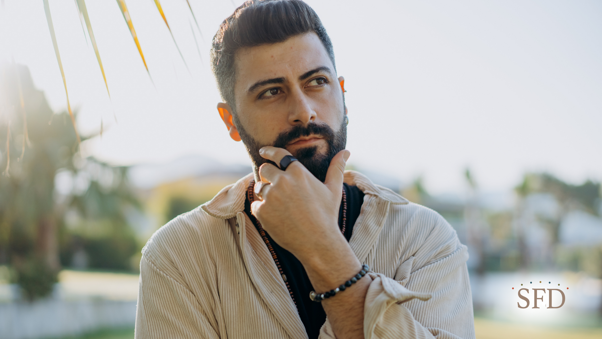 Man with beard, outdoors, thoughtful expression, hand to chin, wearing rings, sunlight.
