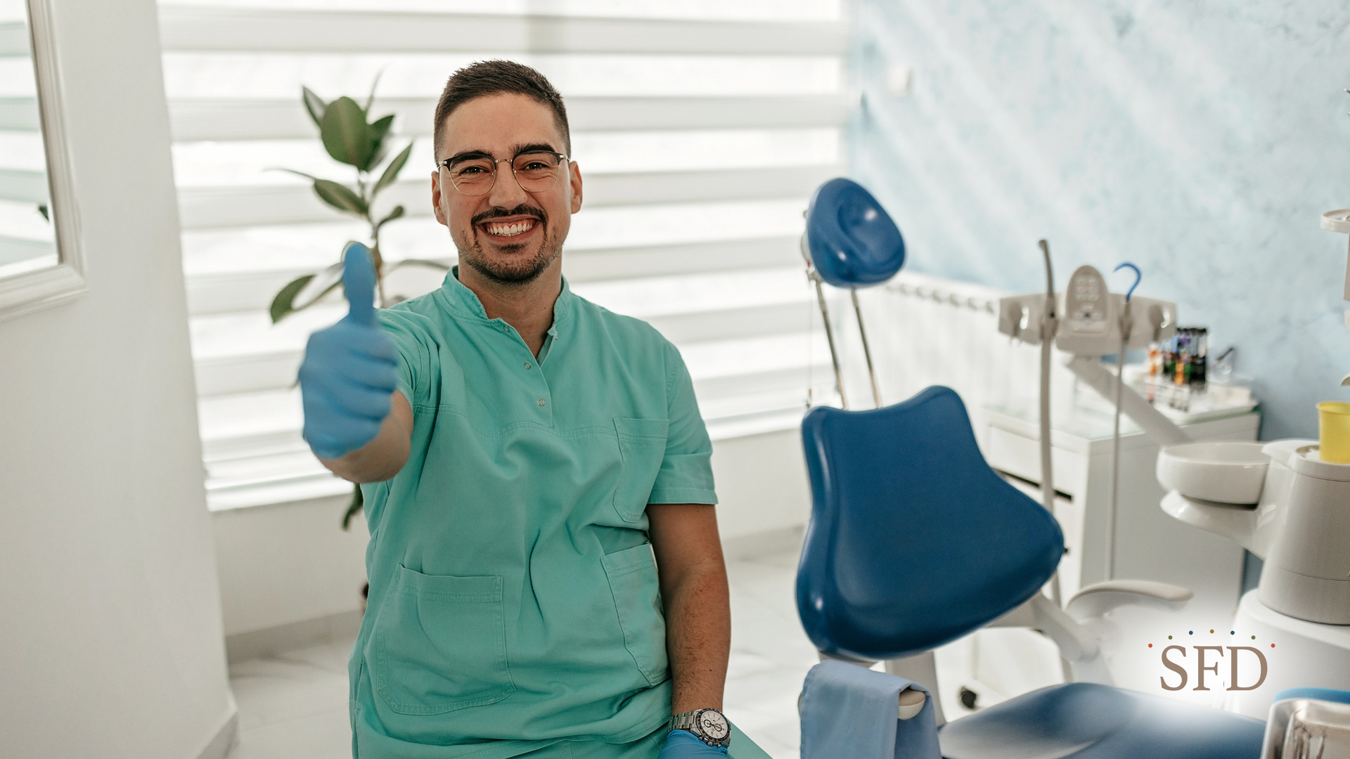 Dentist in teal scrubs giving a thumbs up, in a dental office, smiling.