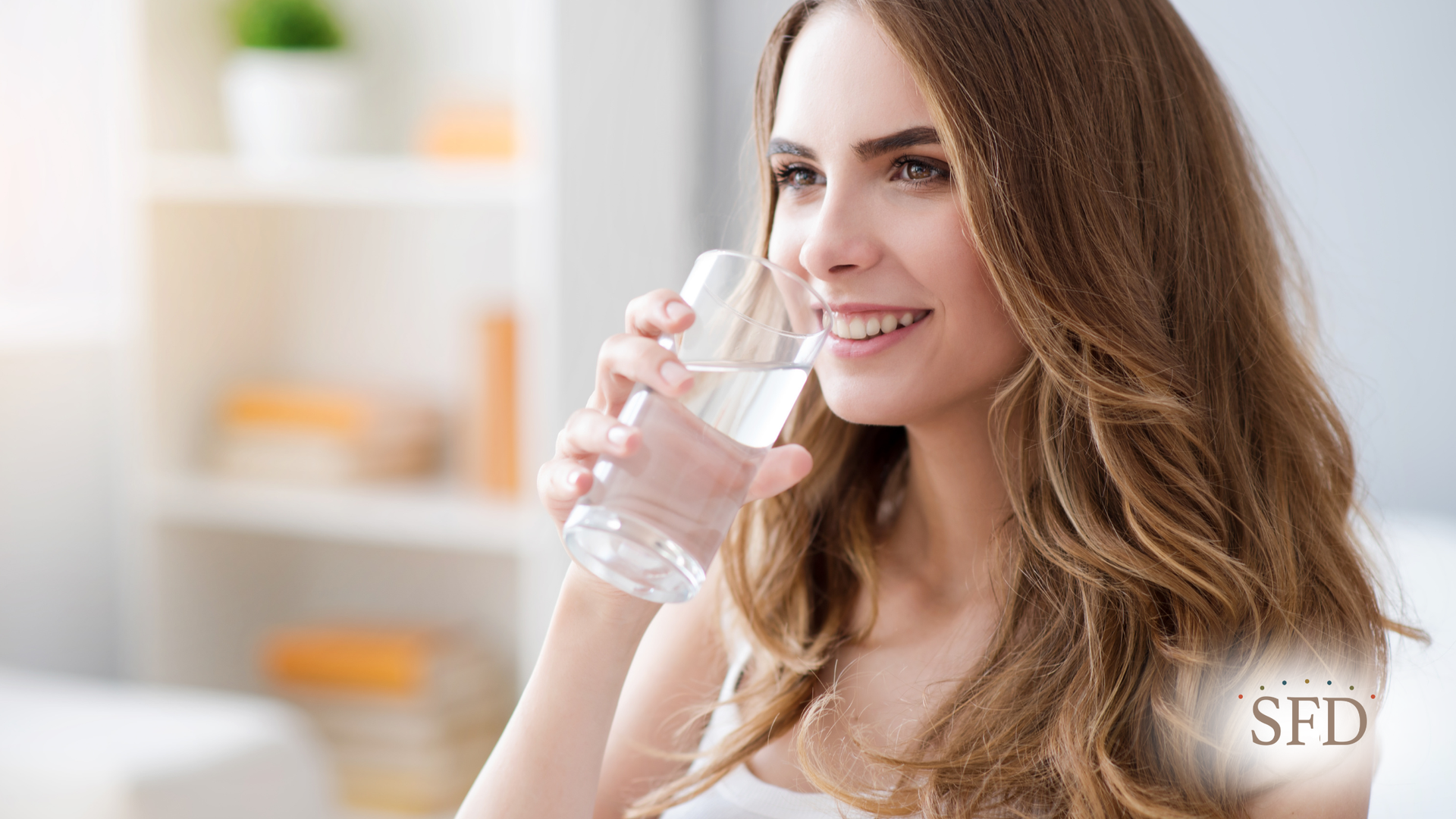 Woman smiles, drinking water from a glass; light-filled room with blurred background.