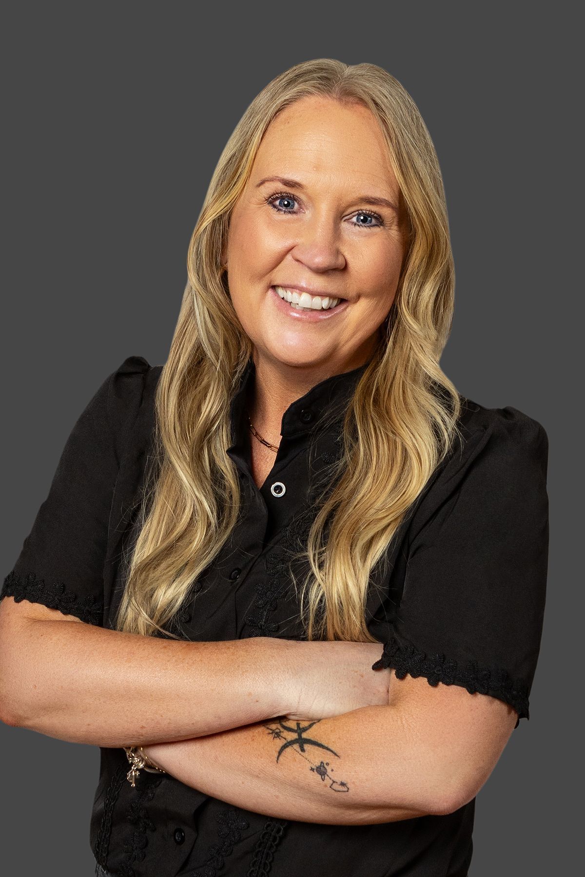 Smiling person with long blonde hair in a black shirt, arms crossed against a gray background