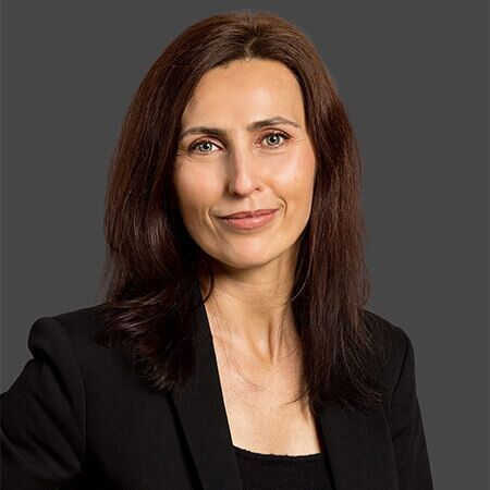 A woman with long brown hair is wearing a black jacket and smiling.