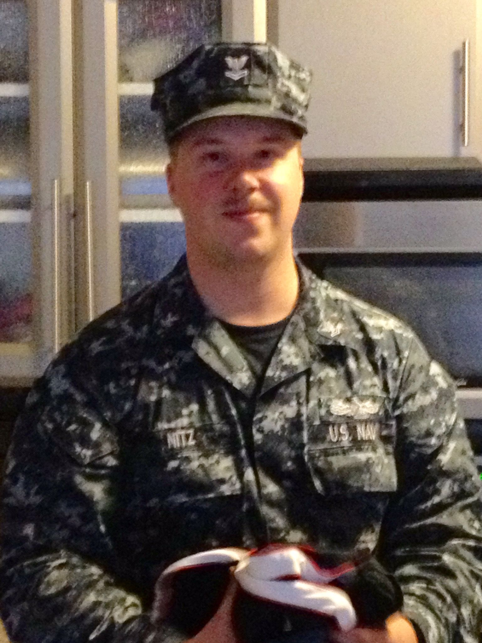 Person in military camouflage uniform and cap, standing indoors and holding an open book.