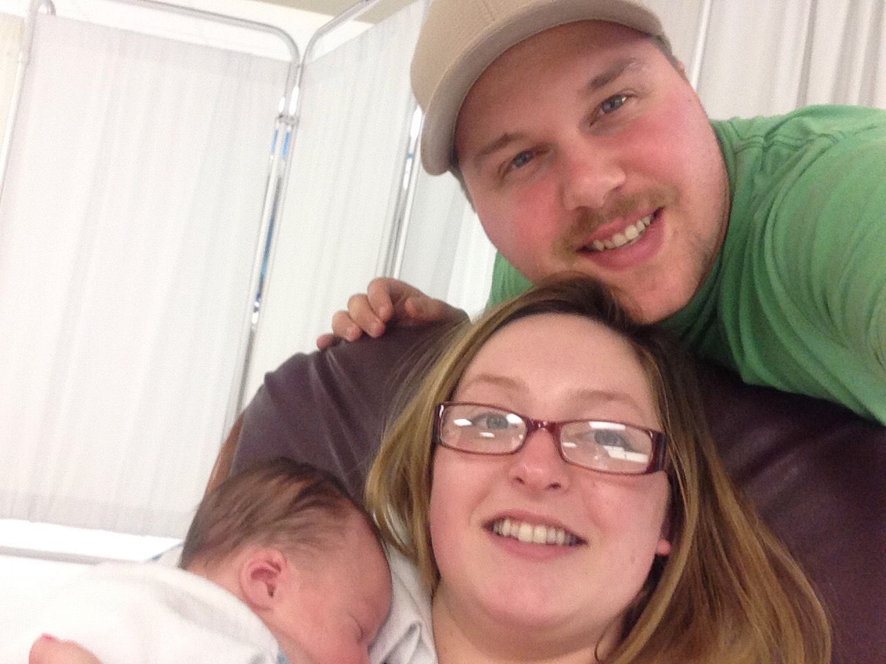 Three people smiling closely together indoors, with a sleeping baby in the foreground