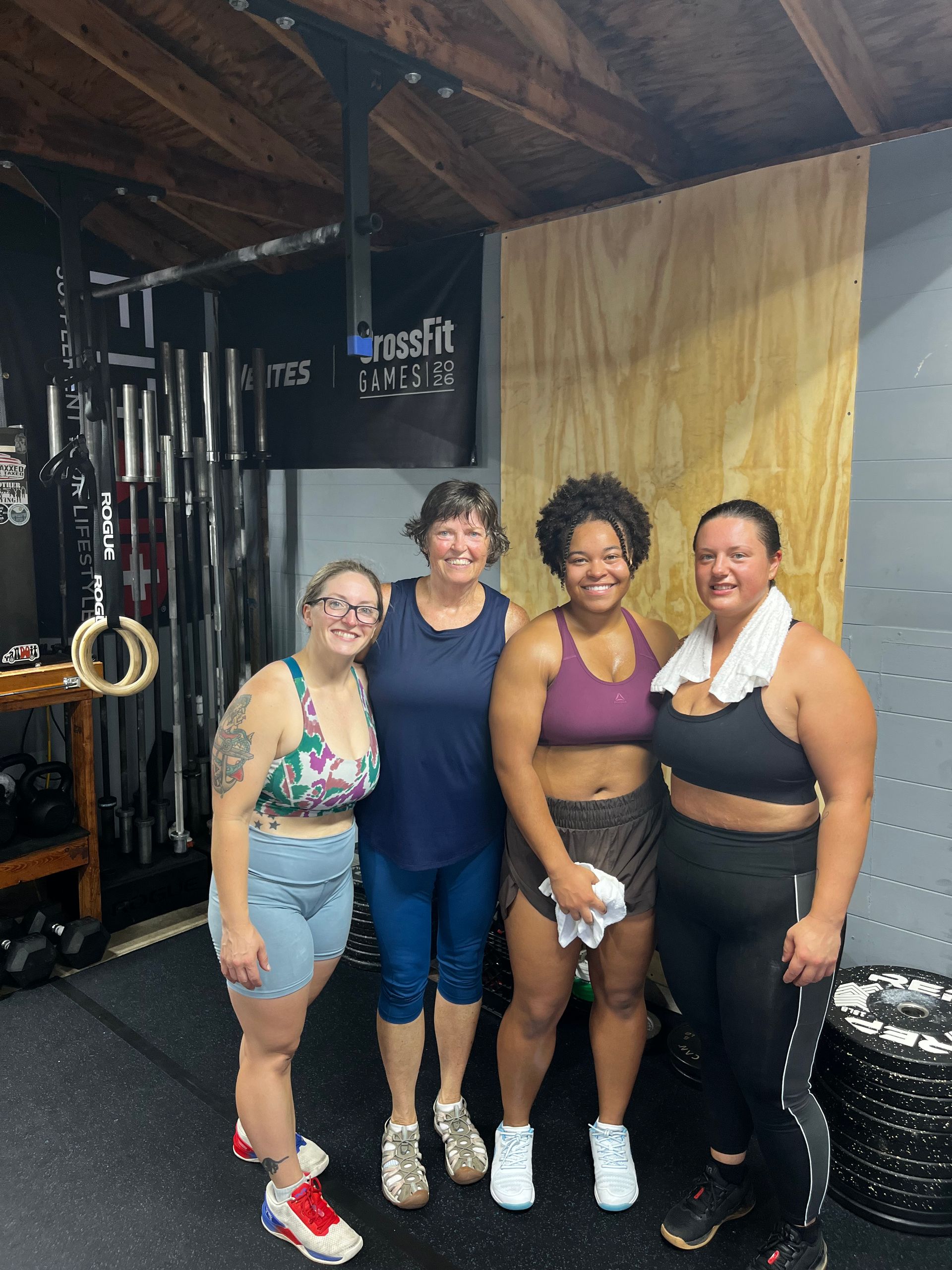 Four women posing together in a gym, smiling in front of workout equipment.