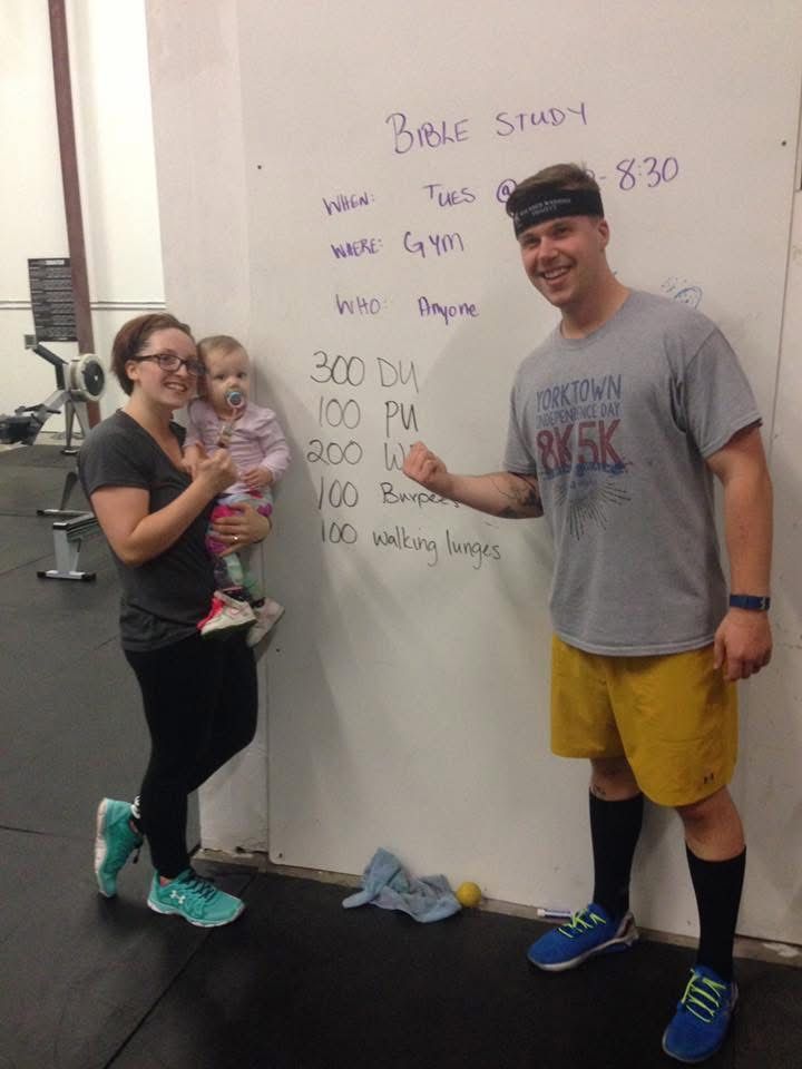 Three people posing by a whiteboard with workout notes in a gym, one holding a baby.