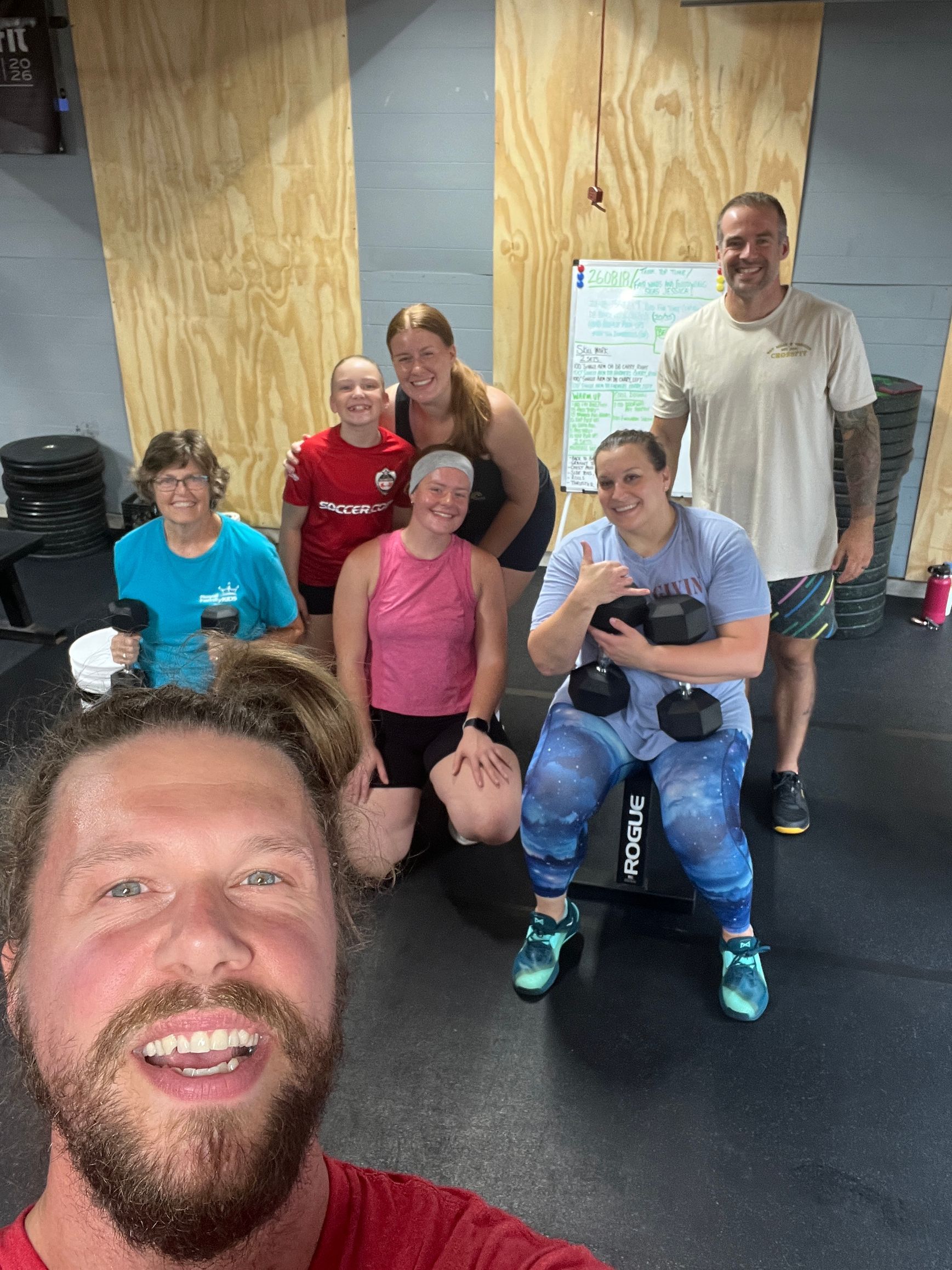 Group of people posing in a gym, with one smiling selfie in the foreground and exercise equipment behind them.