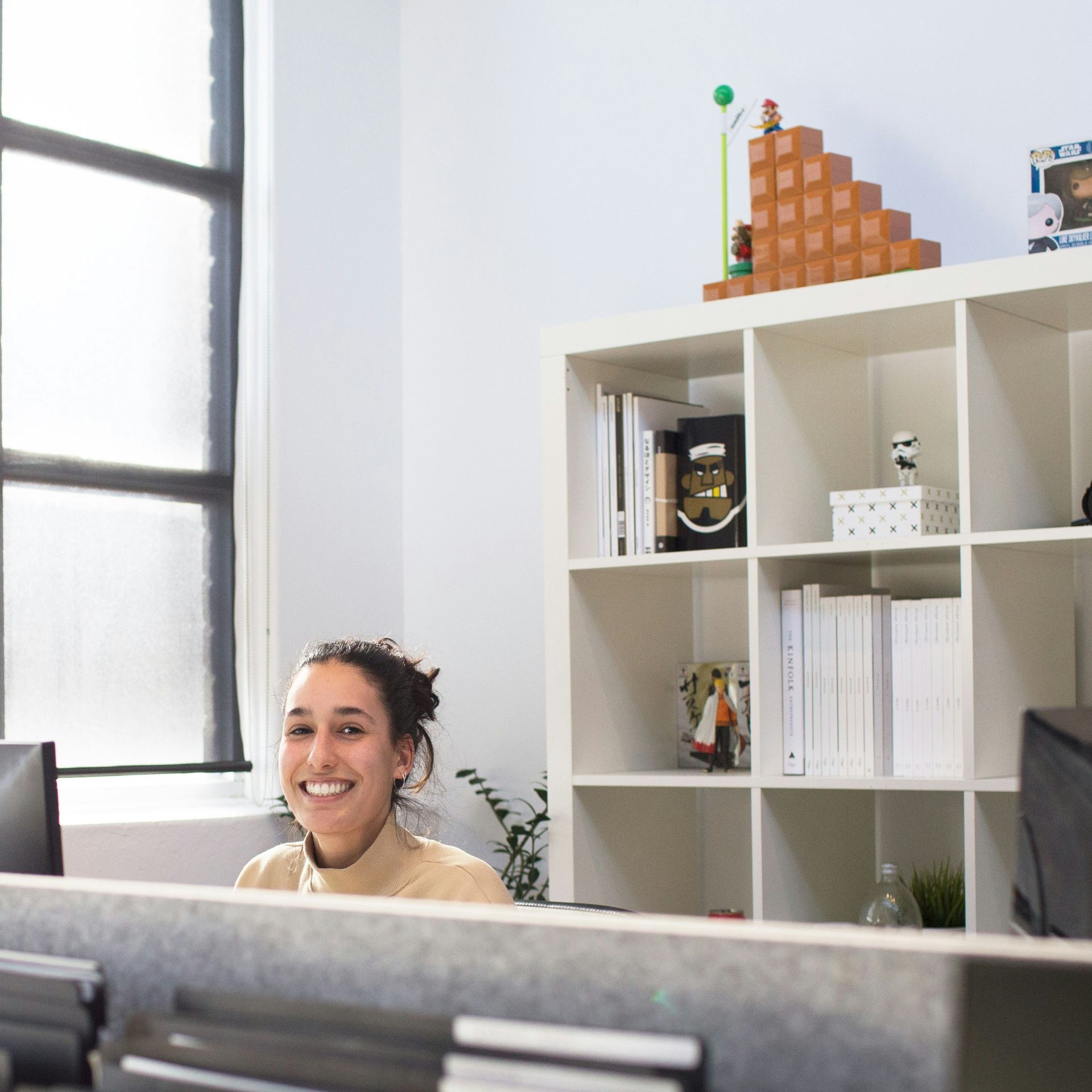 A woman is smiling while sitting at a desk in front of a bookshelf