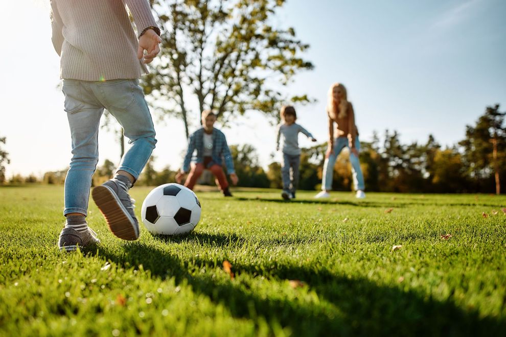 A family plays soccer on a grassy field in the sun, with one person kicking the ball toward three others in the distance.