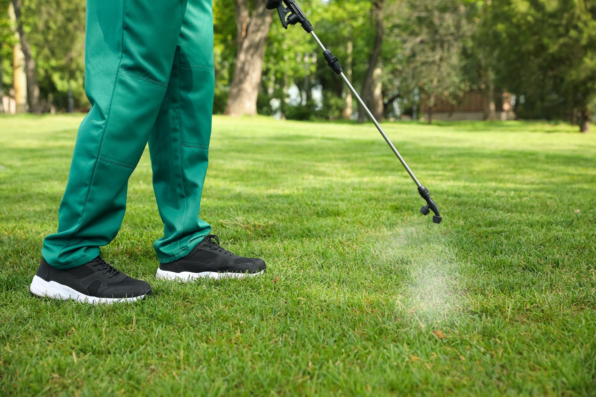 A person in green scrubs uses a spray wand to apply pesticide or fertilizer to a green grassy lawn.