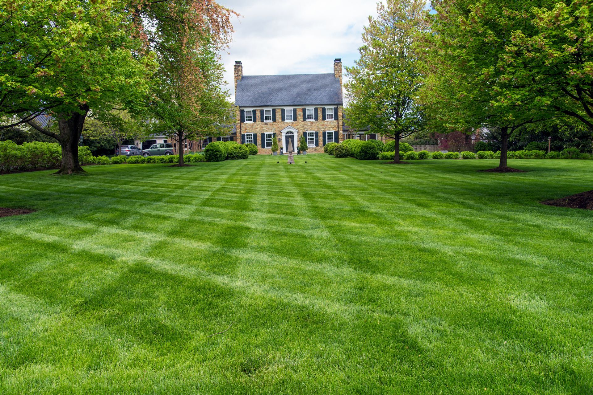 A stately tan brick house stands at the end of a vast, neatly manicured lawn with a cross-hatched pattern of mown grass.